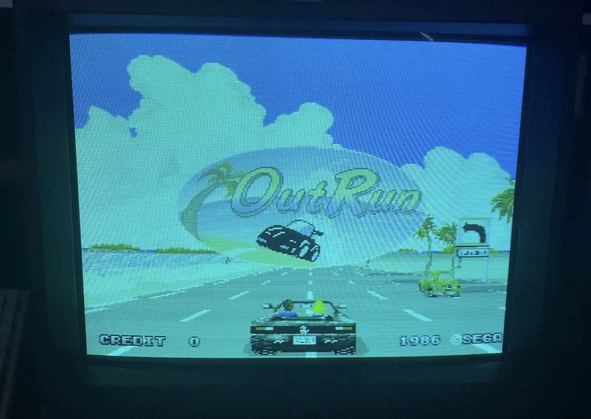 A screenshot of the classic 1986 Sega arcade racing game OutRun, displayed on a CRT monitor. The game shows a pixelated car…