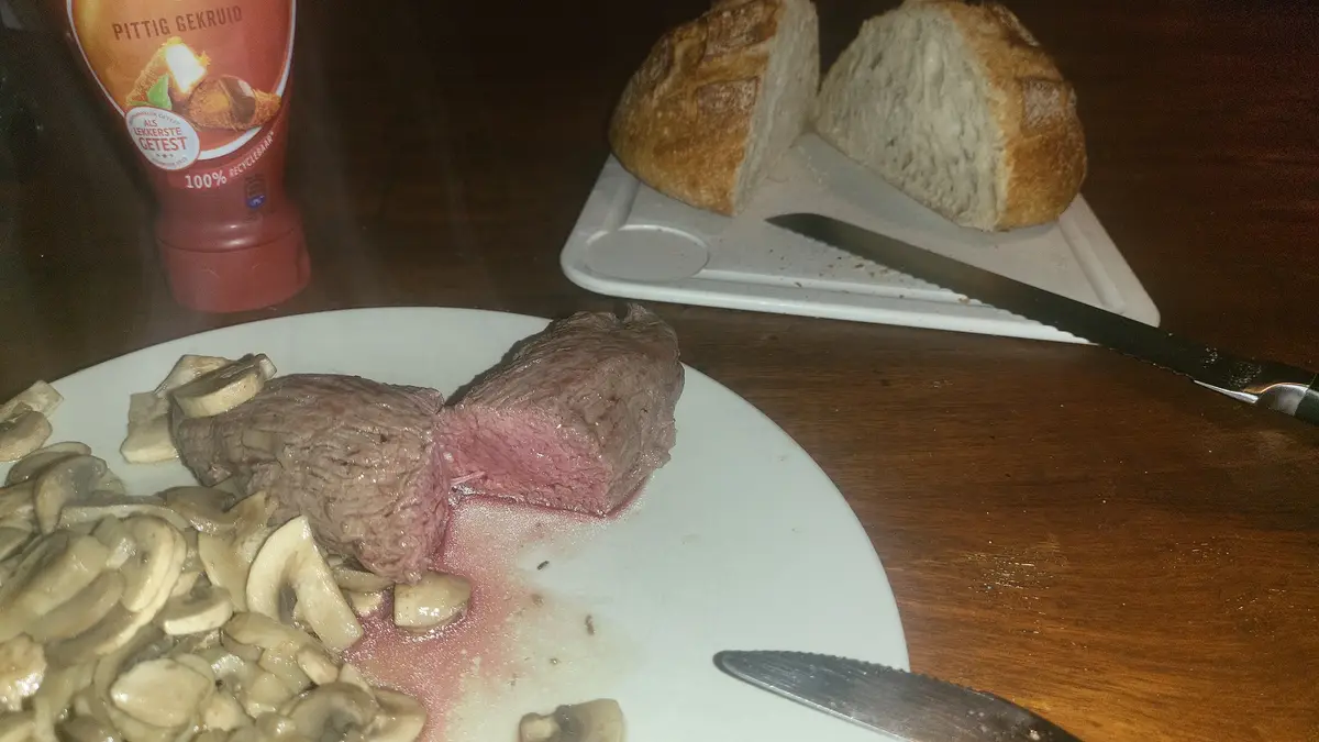 A close-up photo of a sliced medium-rare steak on a white plate, surrounded by sautéed mushrooms, with a loaf of bread and a…