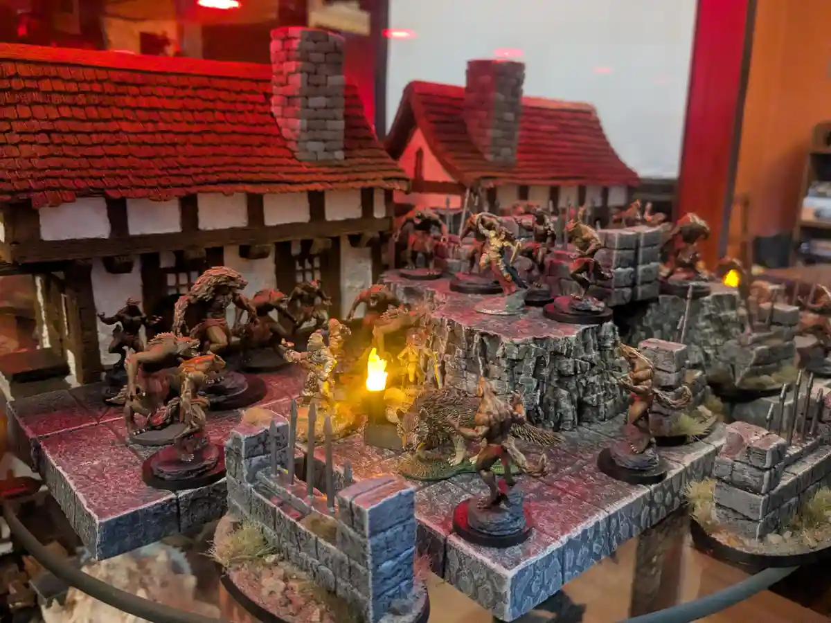 A miniature tabletop wargaming scene depicts a chaotic battle in a medieval village, with numerous painted werewolf figures…