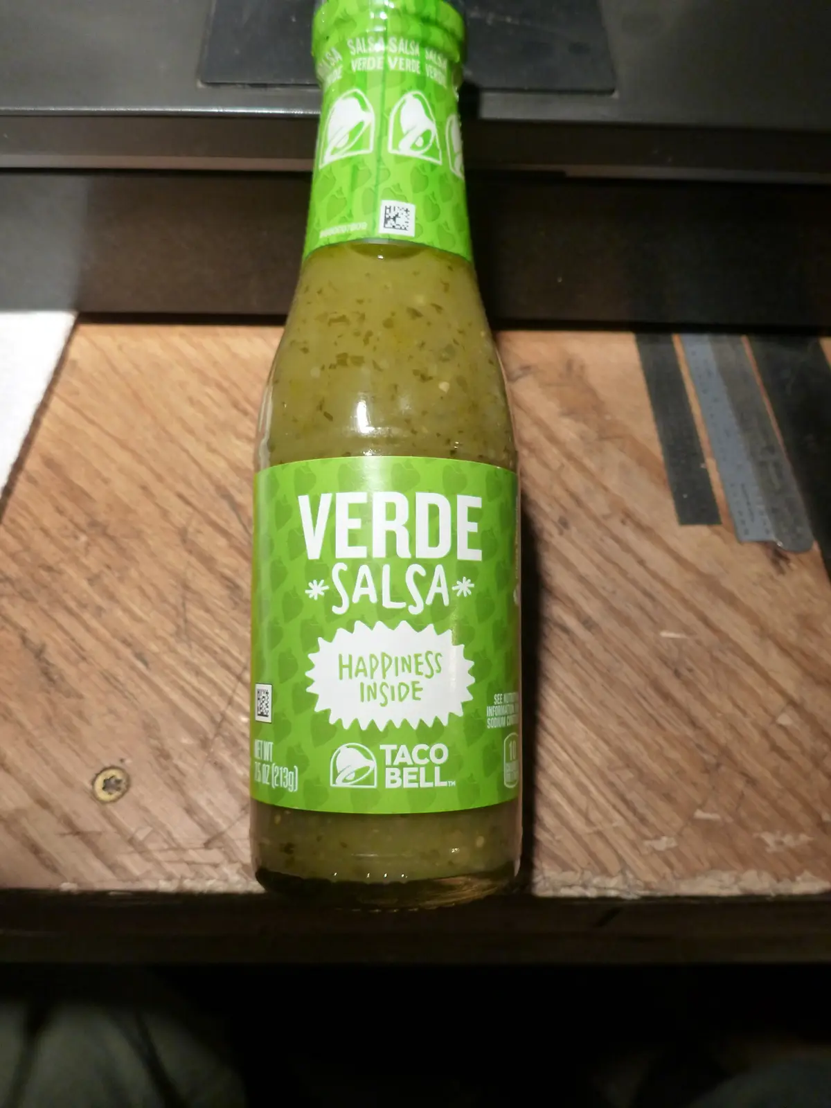 A bottle of Taco Bell Verde Salsa sits on a wooden surface, its green label prominently displaying the brand name and the…