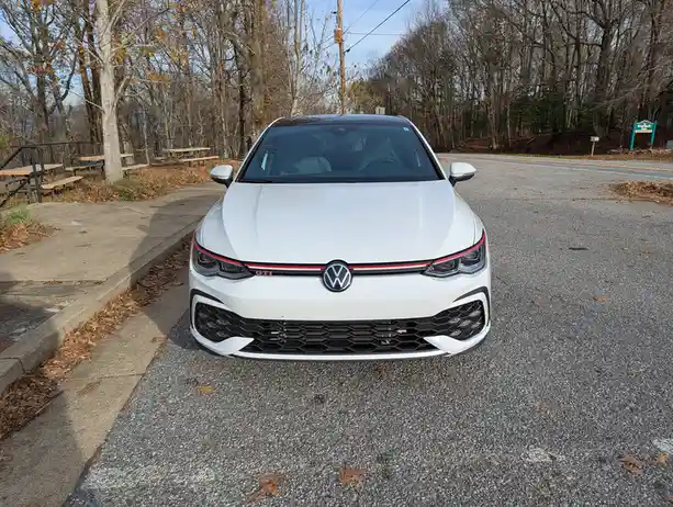 A white Volkswagen GTI is parked on an asphalt road in a wooded area with fallen leaves, facing the camera. The car's front…