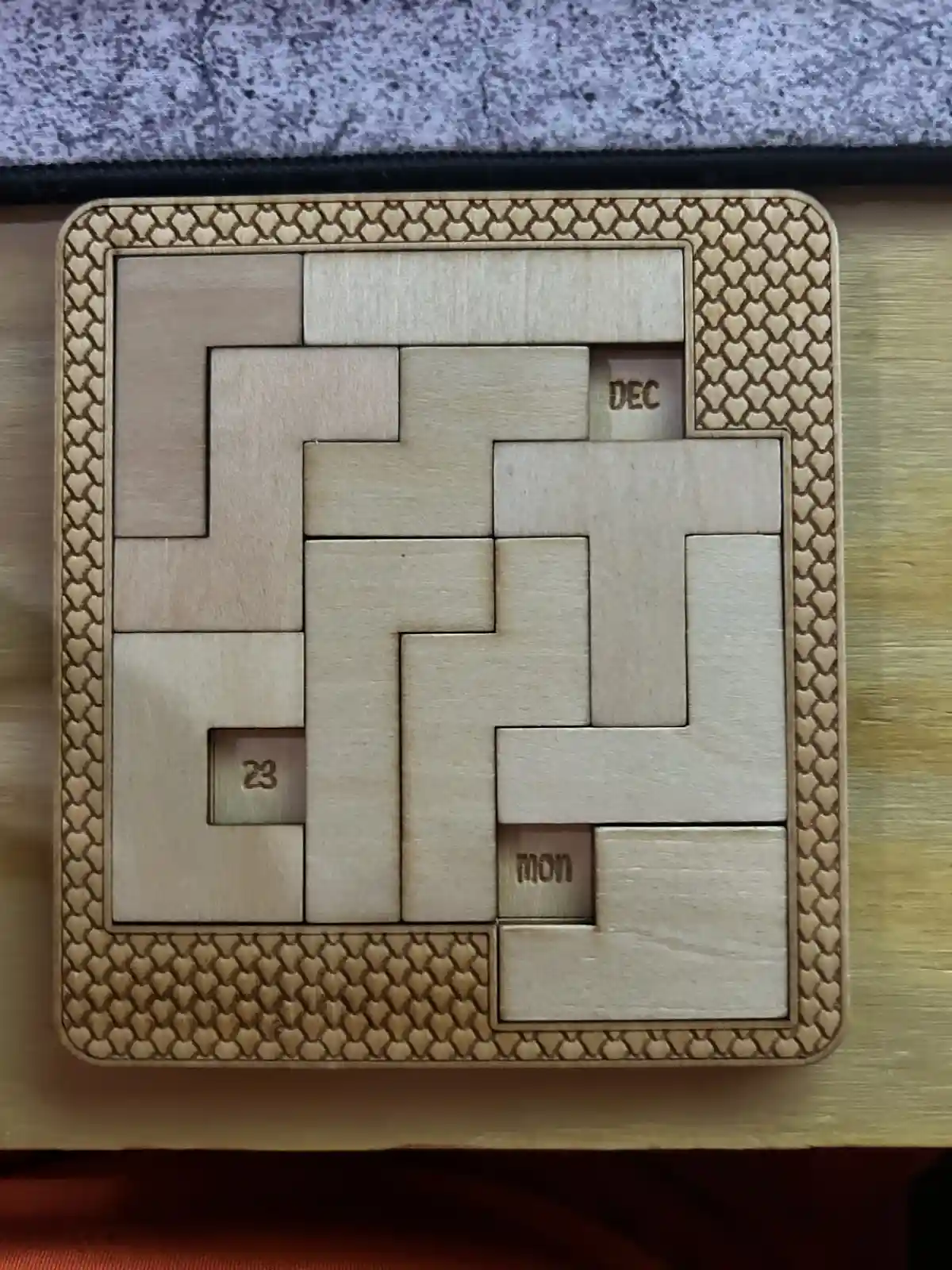A wooden puzzle with interlocking pieces forming a grid, featuring three small recessed squares with engraved text: 'DEC' in…