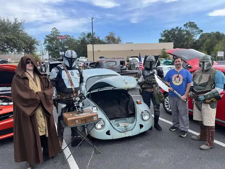 Five men in Star Wars costumes — a Jedi, three Mandalorians, and a man in a blue Star Wars t-shirt — stand around a light…