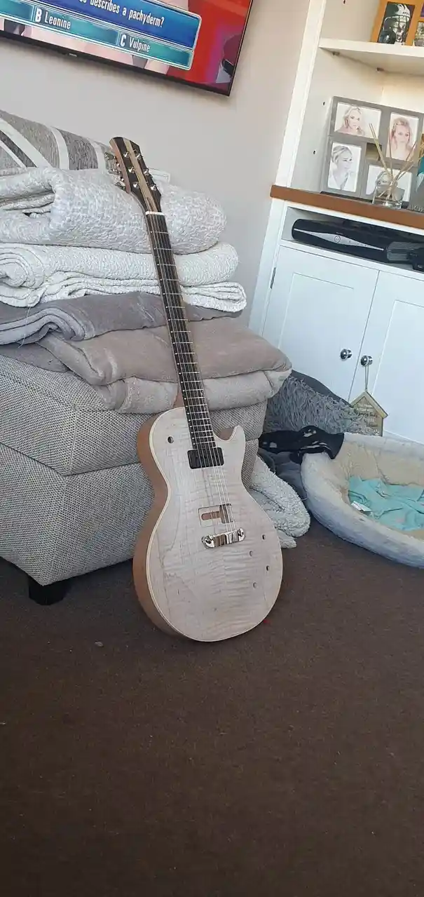 A light-colored electric guitar with a wood grain finish leans against a grey couch piled with blankets in a living room…
