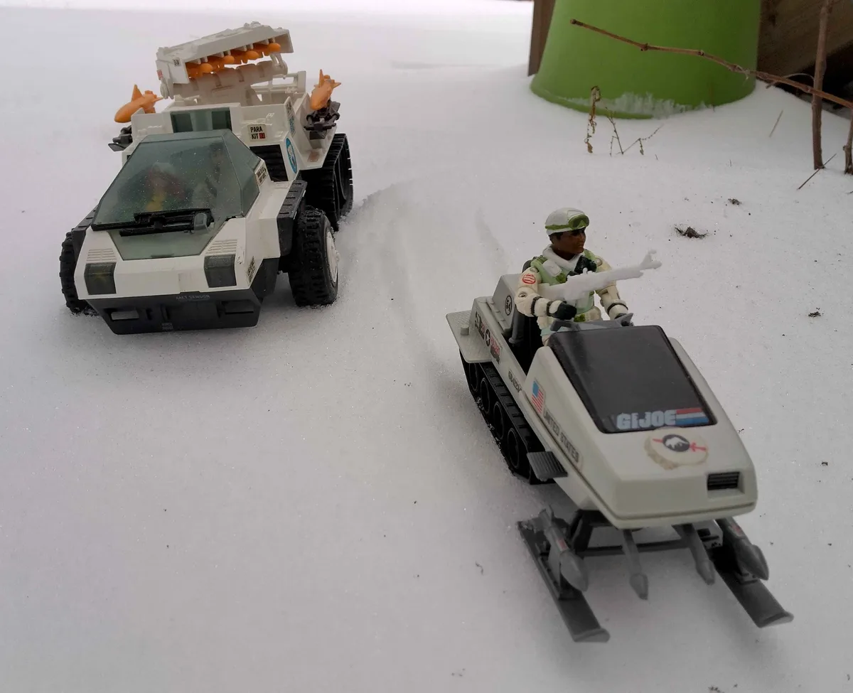 Two G.I. Joe toy vehicles, a snowmobile with a figure and a tracked vehicle, are placed in real snow. The snowmobile has…