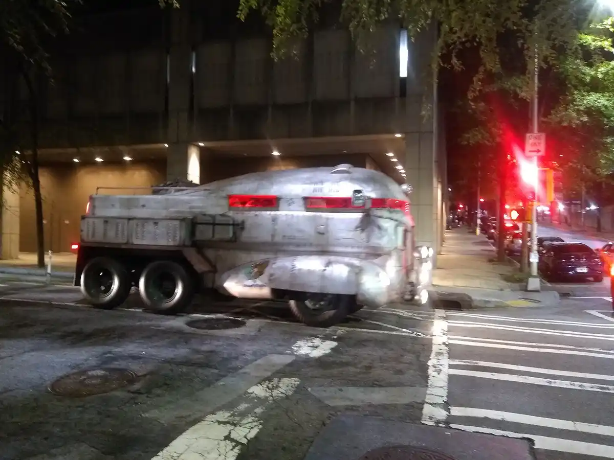 A heavily modified, silver, retro-futuristic truck with a rounded, spaceship-like body is parked at a nighttime city…