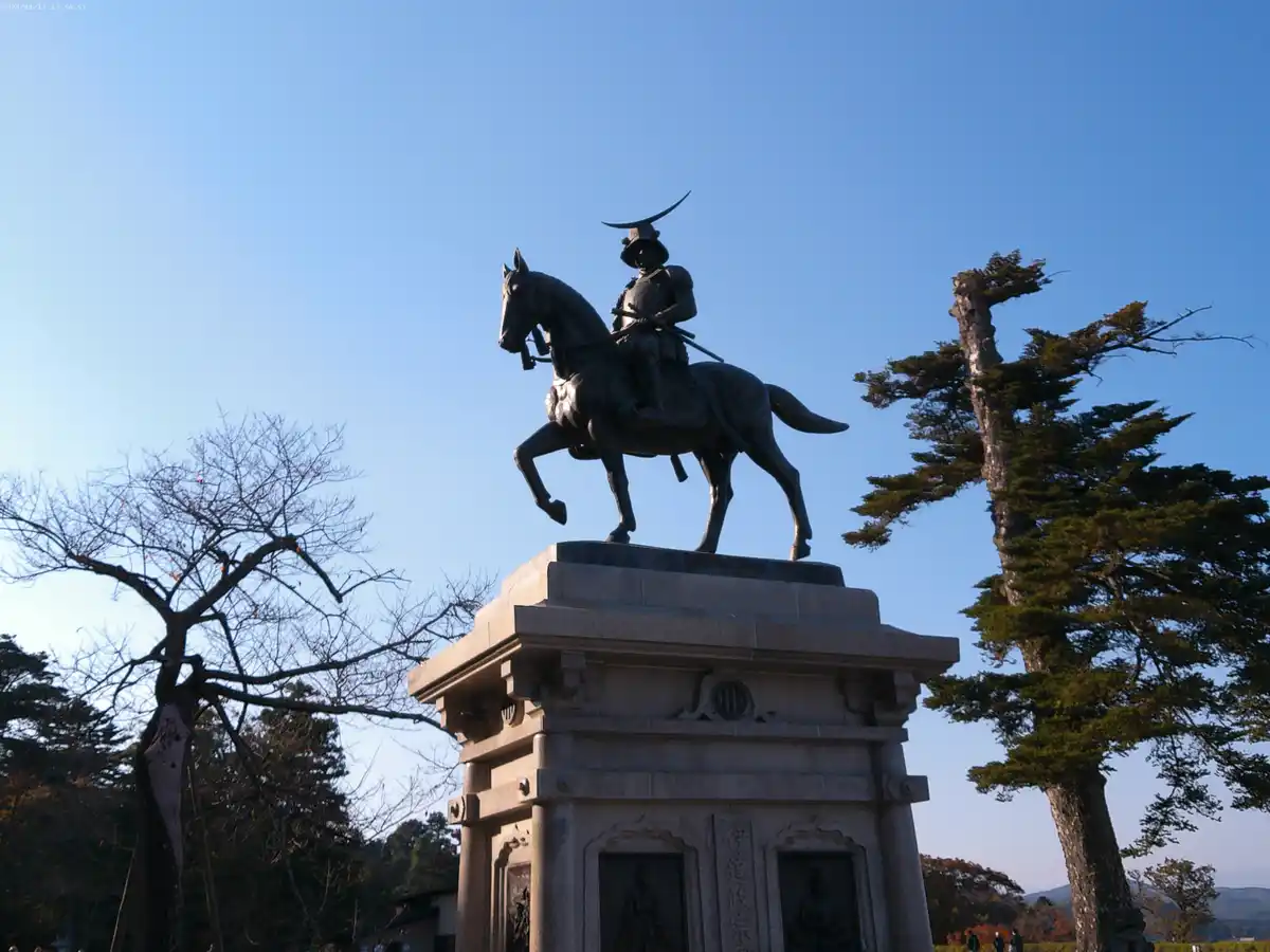 A low-angle photo shows a bronze statue of a samurai on horseback atop a stone pedestal, set against a clear blue sky with…