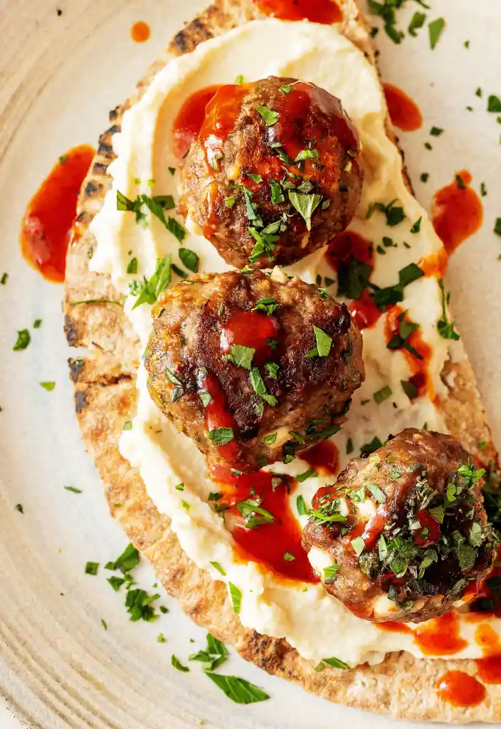 A close-up food photo shows three meatballs resting on a bed of creamy mashed potatoes, which are spread on a piece of…