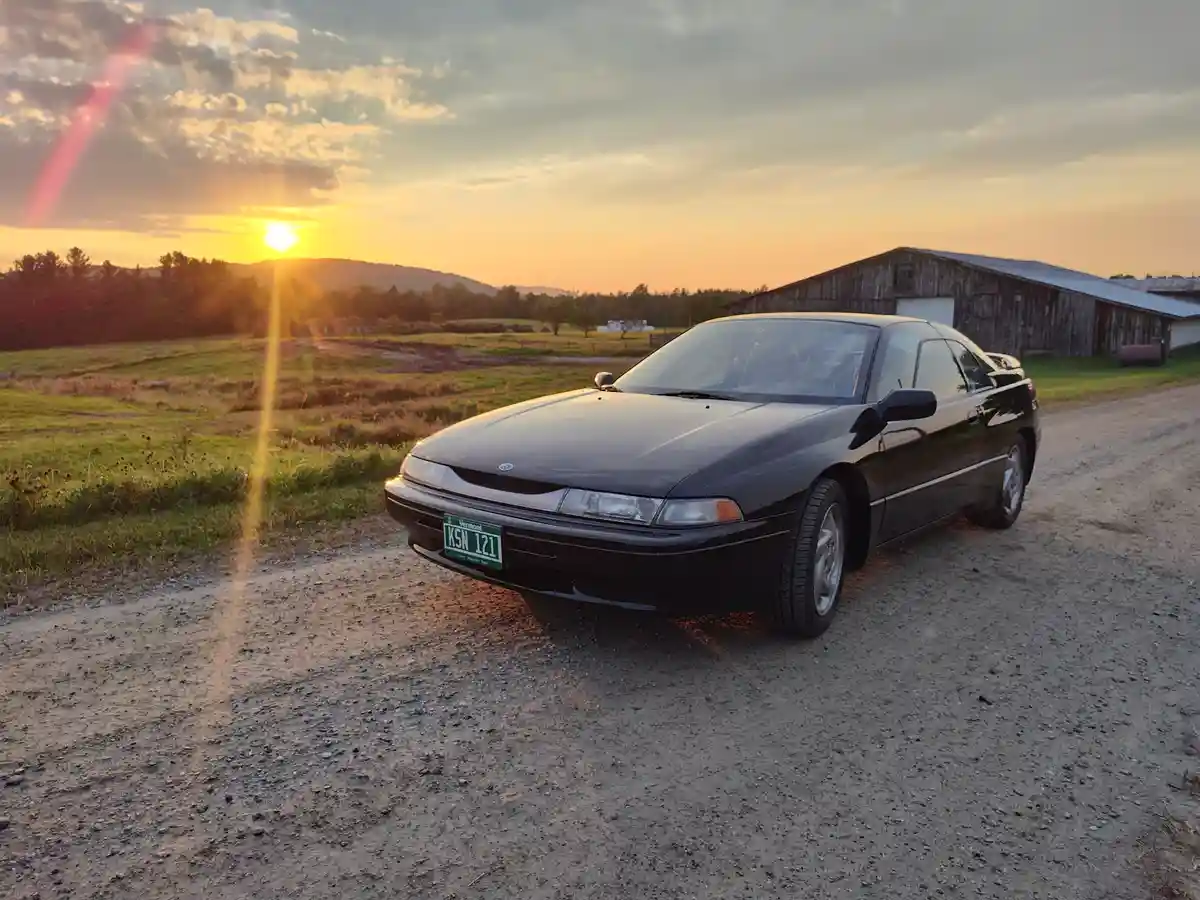 A black 1990s-era sedan, likely a Mazda, is parked on a gravel road at sunset with a rustic barn in the background. The…