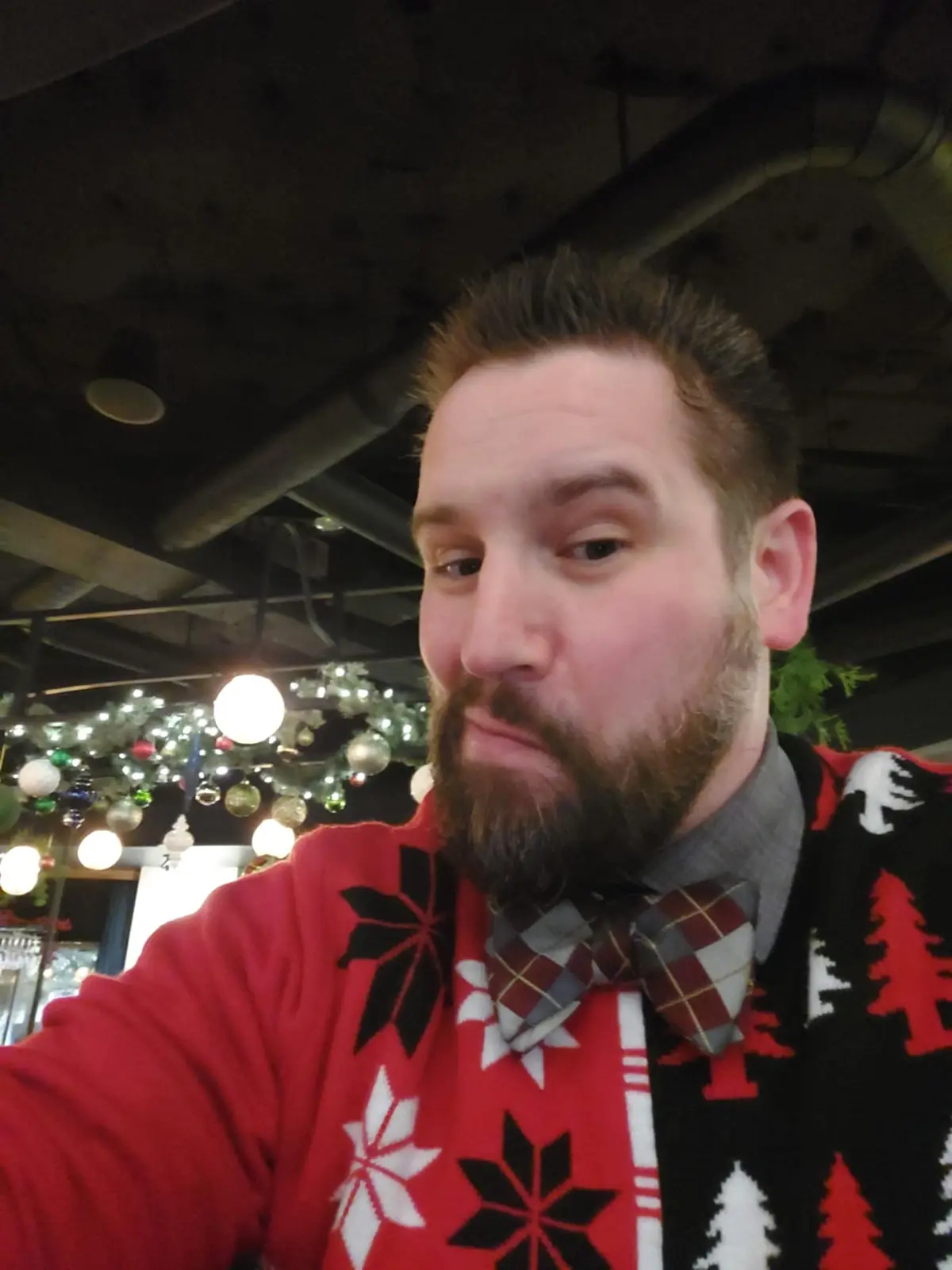 A man with a beard takes a selfie wearing a red, black, and white Christmas sweater with snowflakes and pine trees, paired…