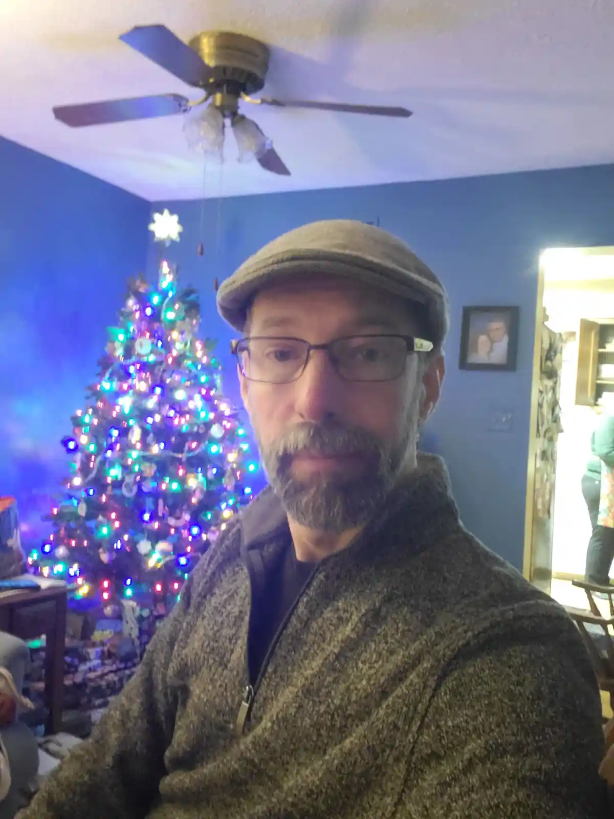 A man with glasses and a flat cap stares directly at the camera with a deadpan expression, standing in front of a brightly…