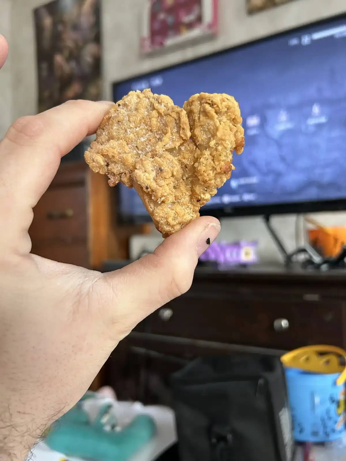 A hand holds up a fried chicken nugget that has broken into two pieces, forming a heart shape. In the background, a TV…