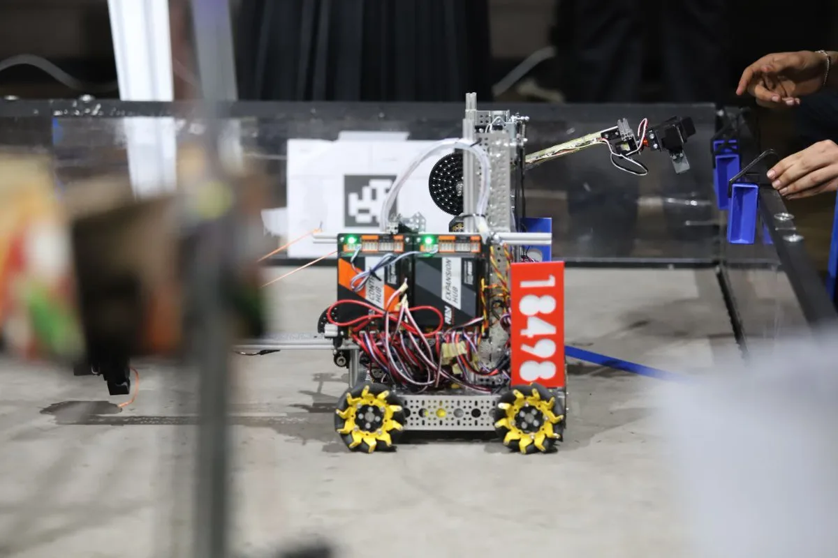 A small, wheeled robot with exposed wiring, yellow spiked wheels, and a red number plate reading '18498' is shown on a…