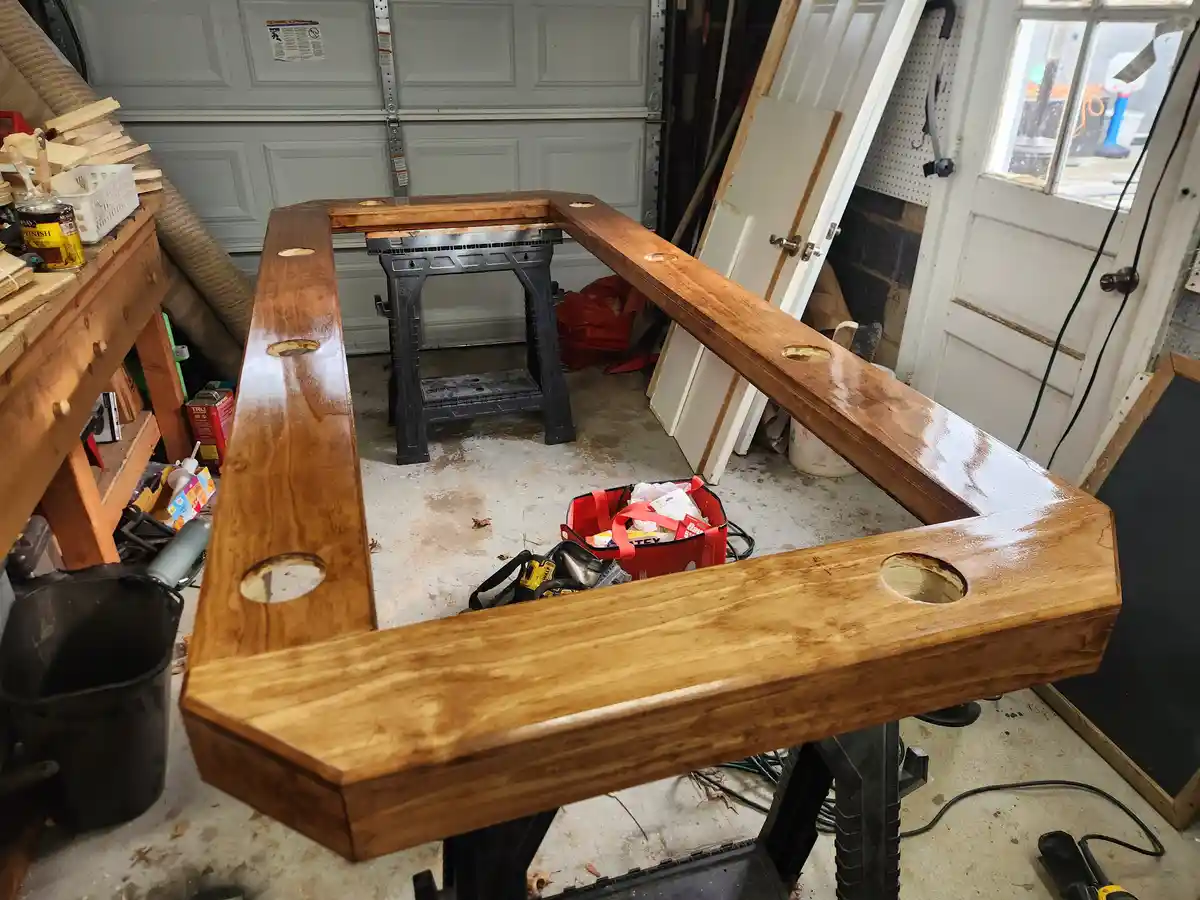 A large, L-shaped wooden table frame sits on sawhorses in a cluttered garage workshop, featuring circular holes drilled into…