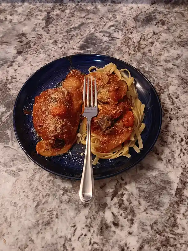 A top-down photo of a blue plate on a speckled countertop, holding pasta with a meaty sauce and a large piece of chicken…