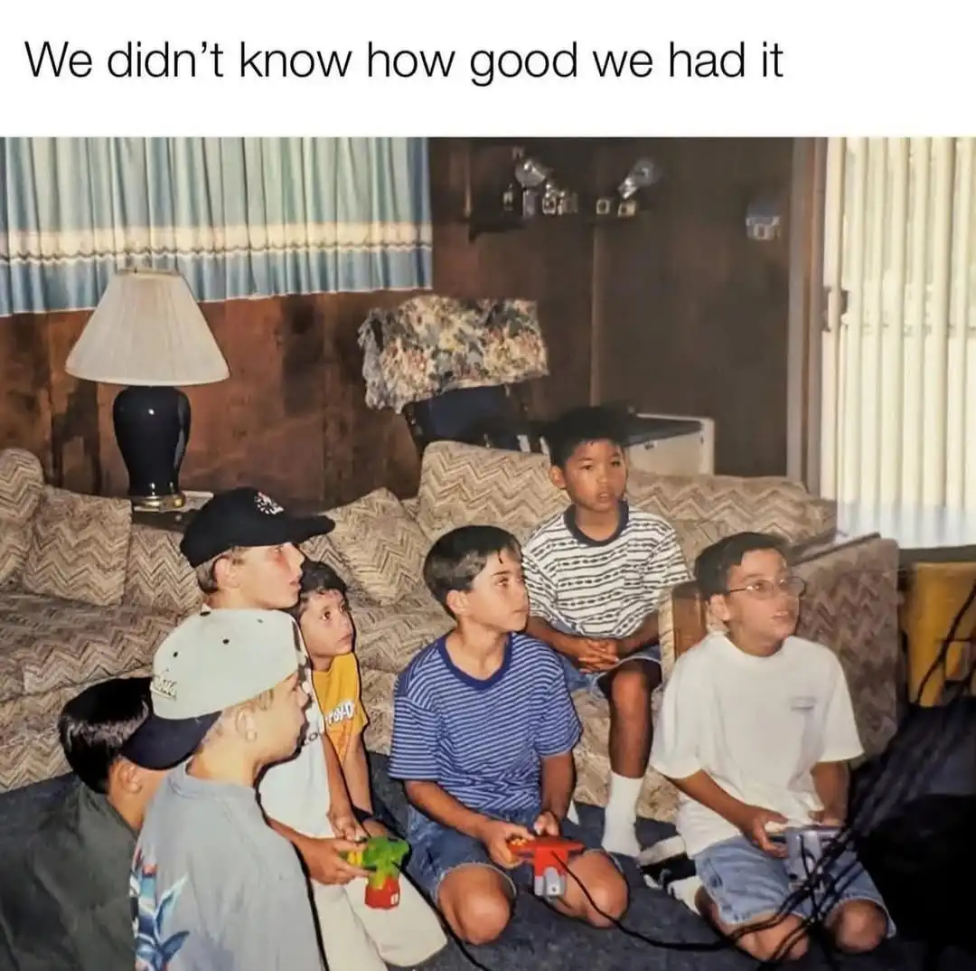A vintage photo shows seven young boys sitting on a patterned couch and floor in a living room, intensely focused on playing…