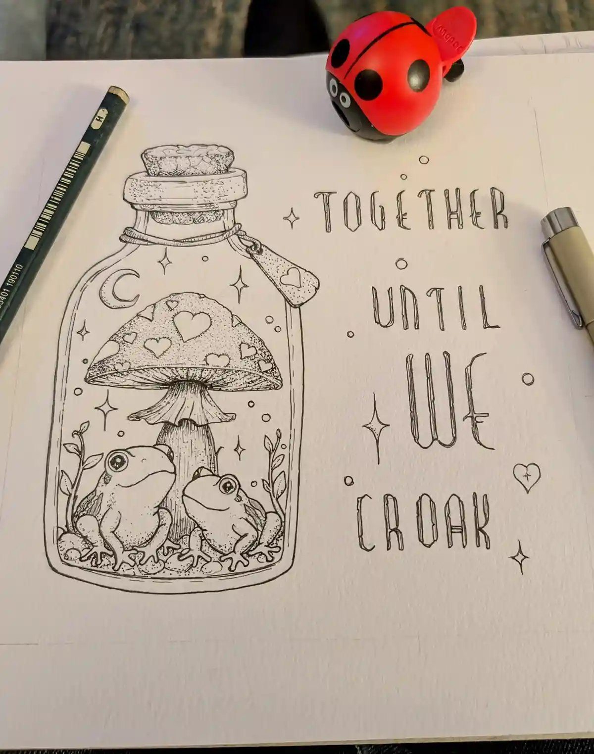 A black-and-white line drawing on paper shows two frogs sitting under a mushroom inside a glass bottle with a cork stopper…
