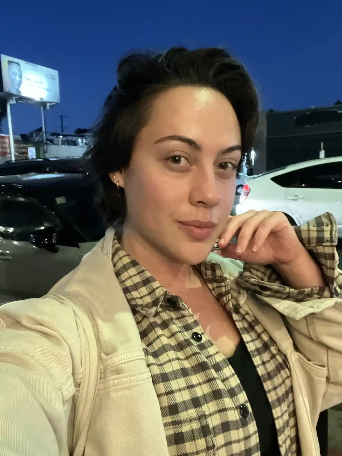 A selfie of a person with short dark hair, wearing a beige jacket over a plaid shirt, posing with their hand near their chin…