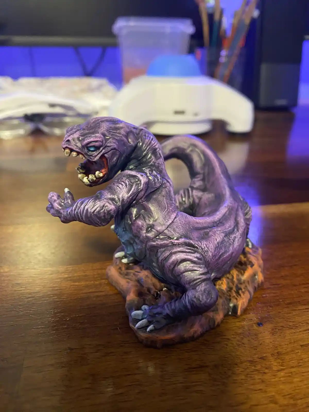 A purple, scaly monster figurine with sharp teeth and claws is posed aggressively on a wooden table. The figurine has one…