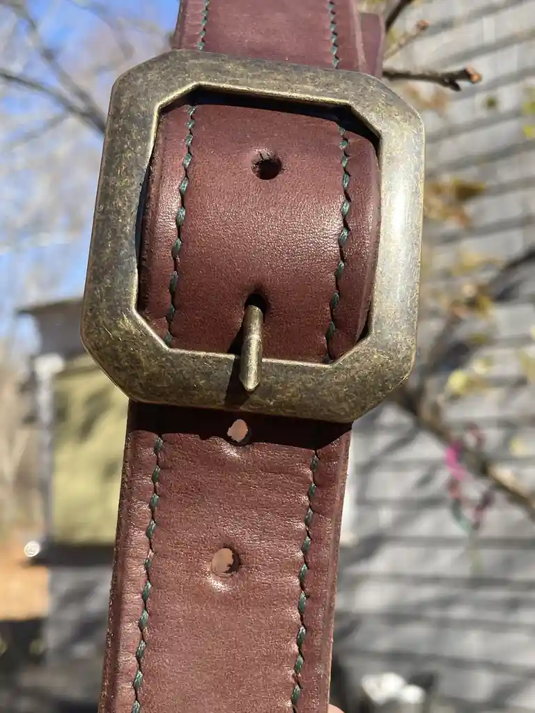 A close-up photo of a brown leather belt with a tarnished brass buckle, showing multiple holes worn through the leather…