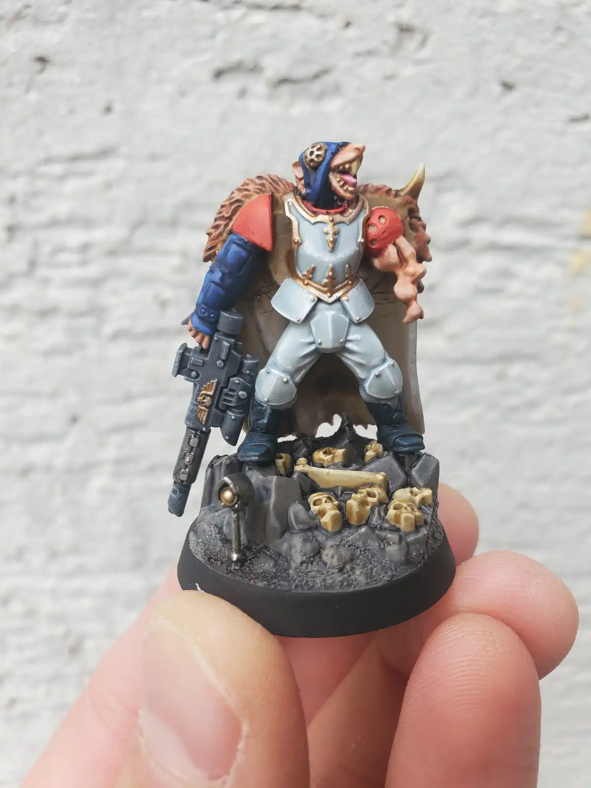 A hand holds a painted miniature of a fantasy warrior with a blue hood, silver armor, and a red shoulder pad, wielding a…