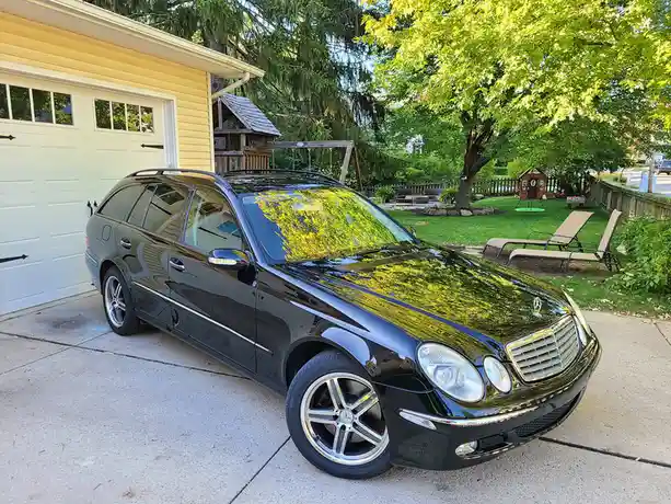 A glossy black Mercedes-Benz E-Class station wagon is parked in a driveway next to a garage, with a lush green backyard…