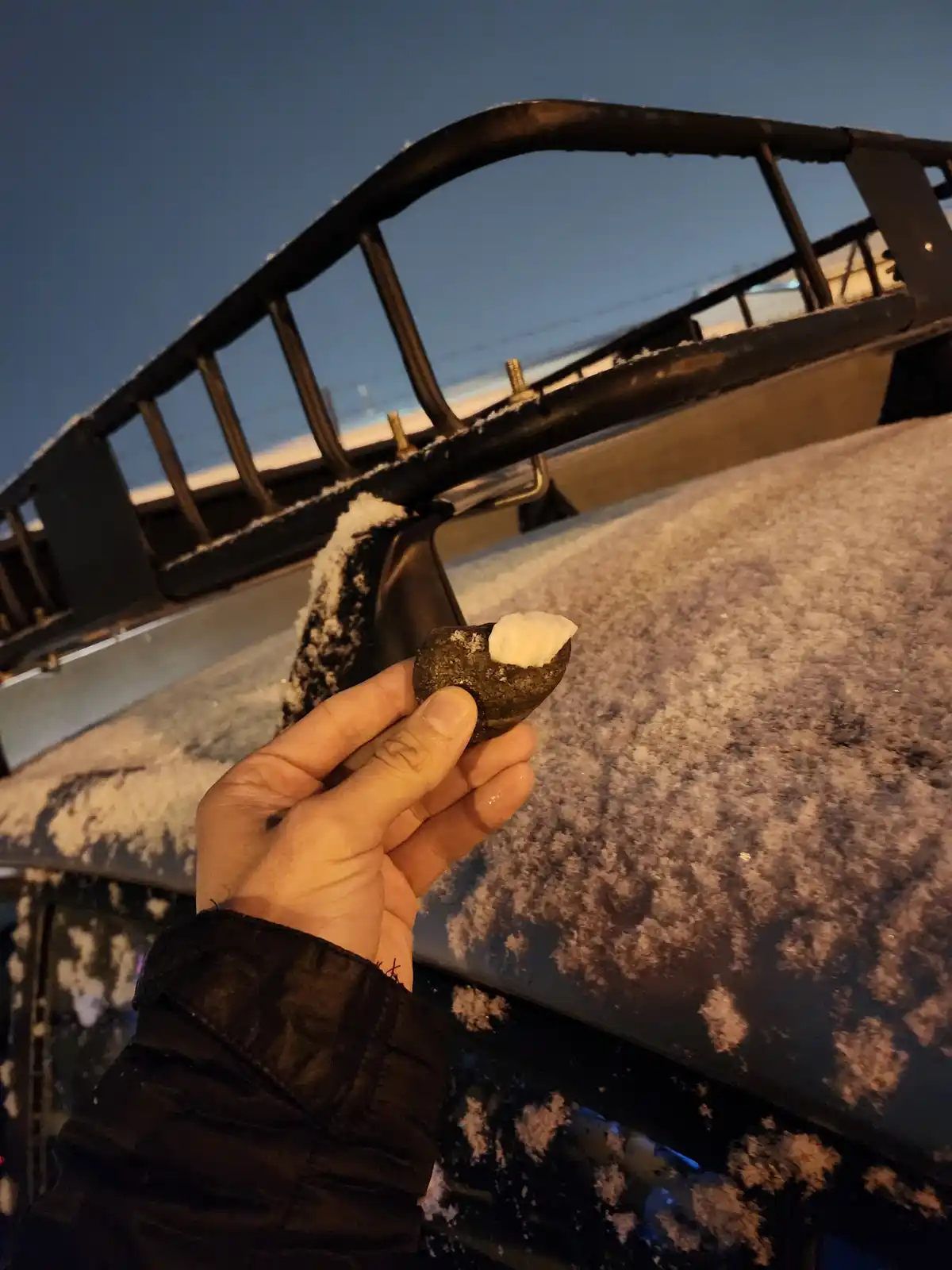 A hand holds a small, dark rock with a scoop of snow embedded in it, in front of a snow-covered car roof and a black roof…