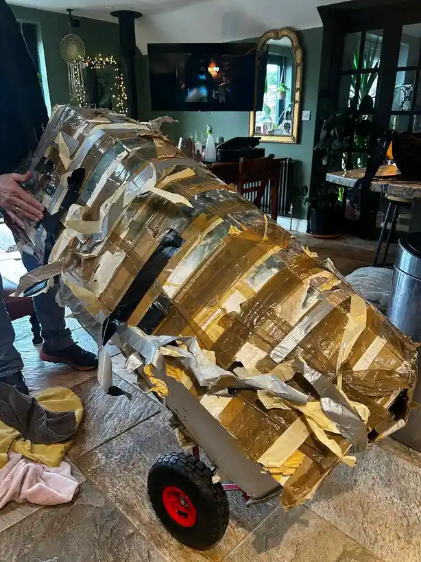 A person is holding a large, homemade vehicle constructed from cardboard and covered extensively with strips of duct tape…