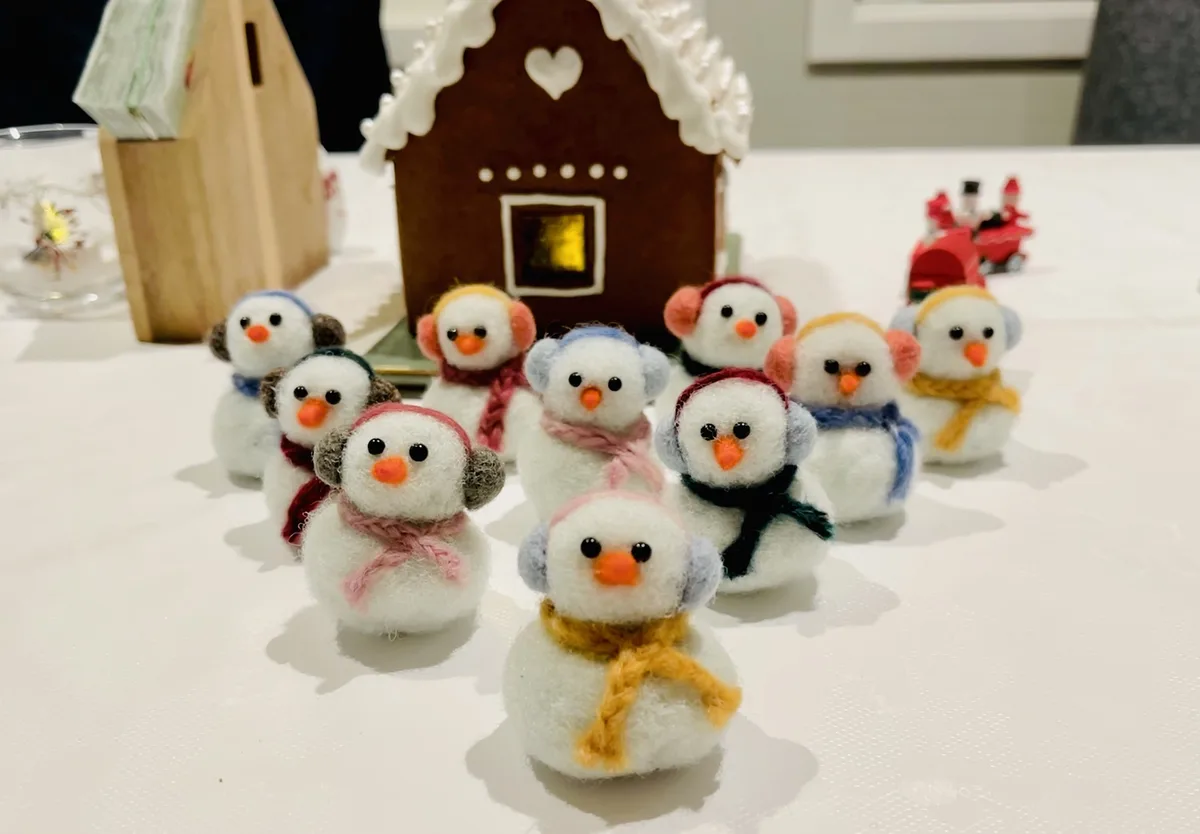 A group of small, handmade felt snowman figurines, each wearing colorful scarves and earmuffs, are arranged on a white table…