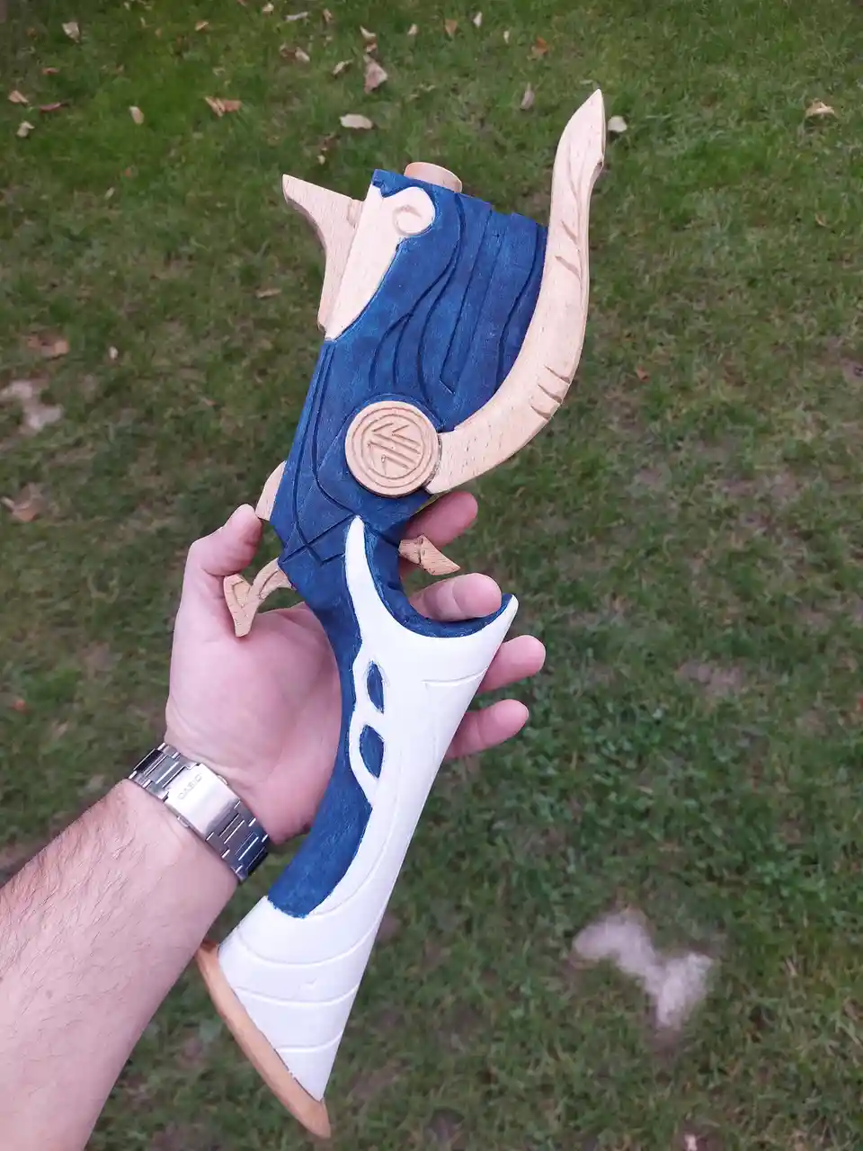 A hand holds a custom wooden replica of the 'Kali' weapon from the game 'Genshin Impact', painted blue and white with carved…