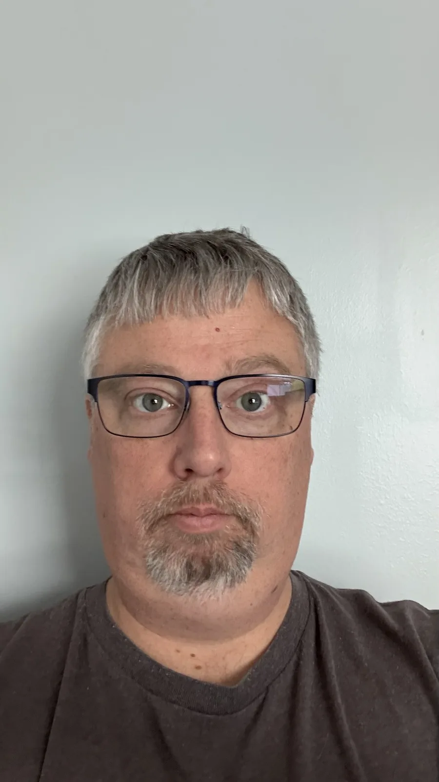 A man with short gray hair, glasses, and a goatee stares directly at the camera with a deadpan expression against a plain…