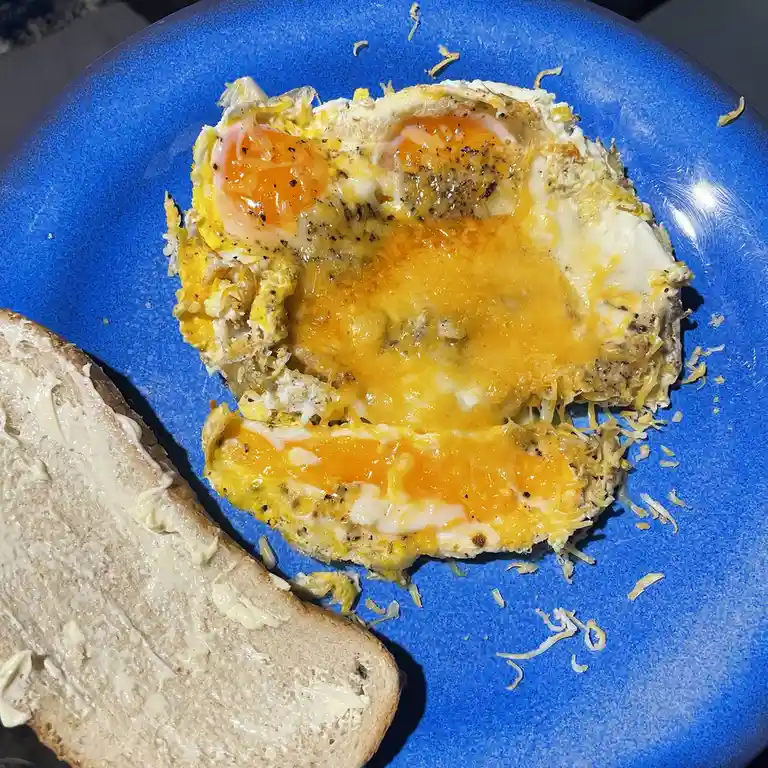 A close-up photo shows a blue plate with a fried egg, melted cheese, and a slice of buttered bread. The egg is split open…