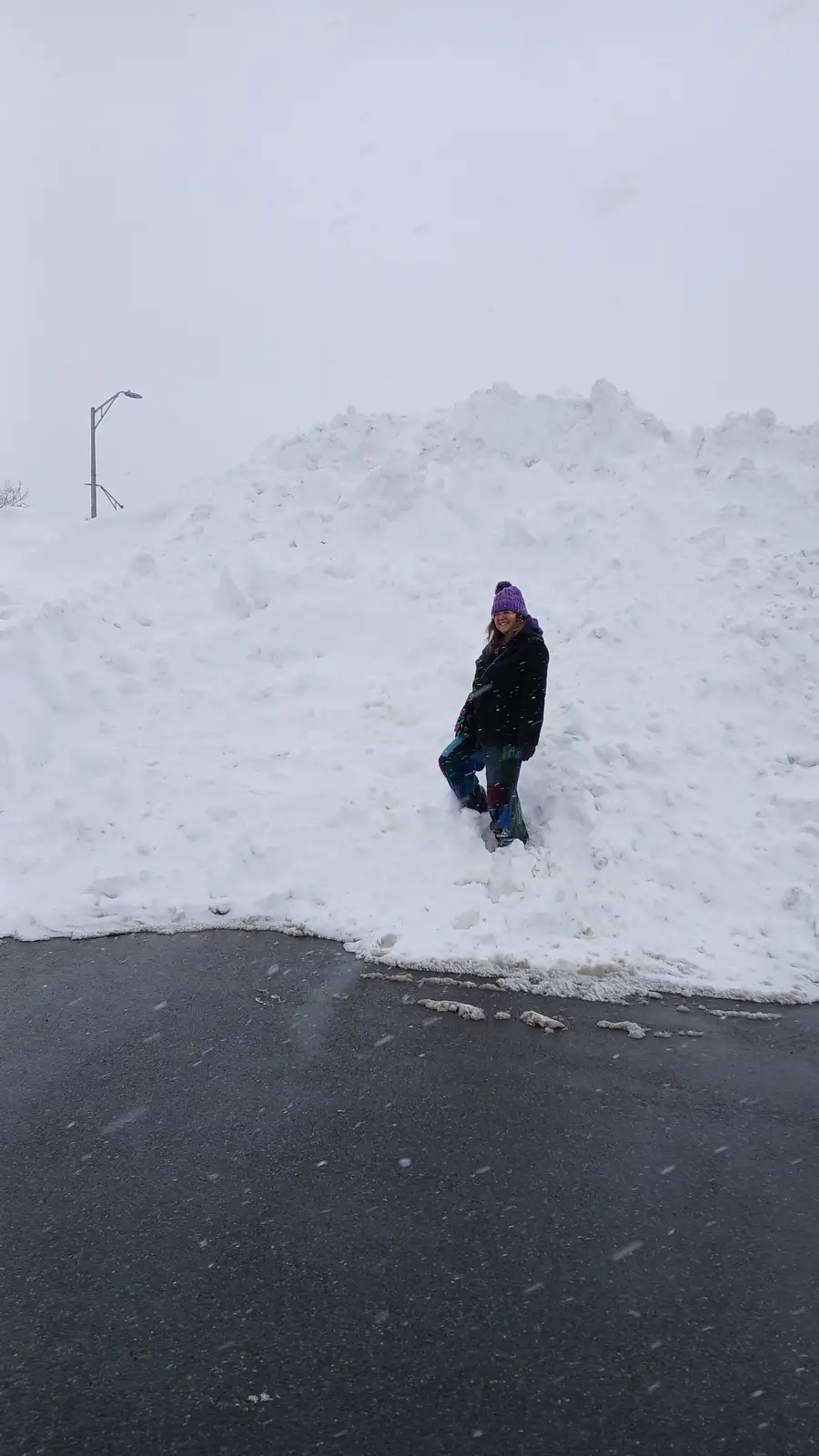 A person wearing a purple beanie, glasses, a black coat, and colorful pants stands smiling in front of a large snowbank on a…