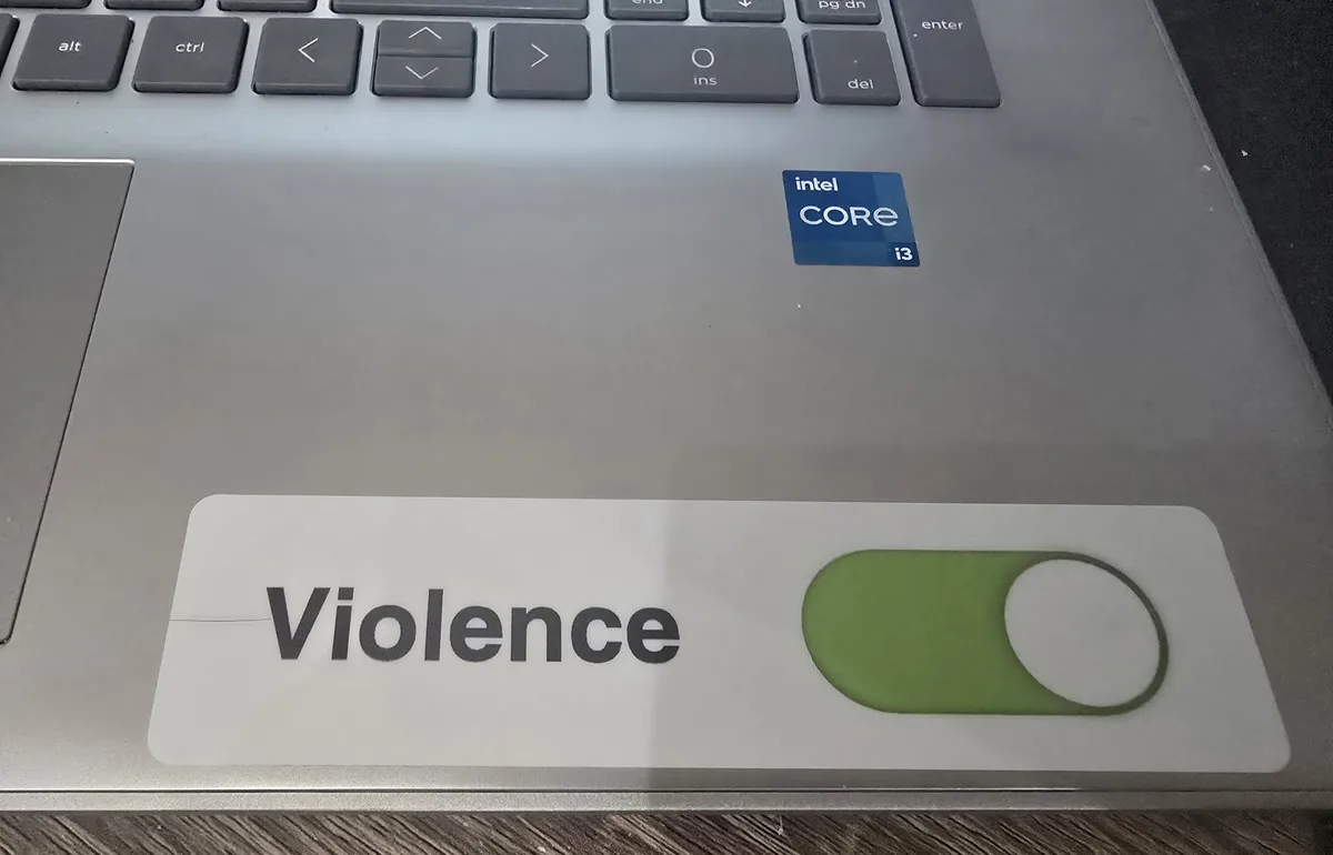 A close-up photo of a laptop's palm rest area, showing a sticker with the word 'Violence' next to a green toggle switch…