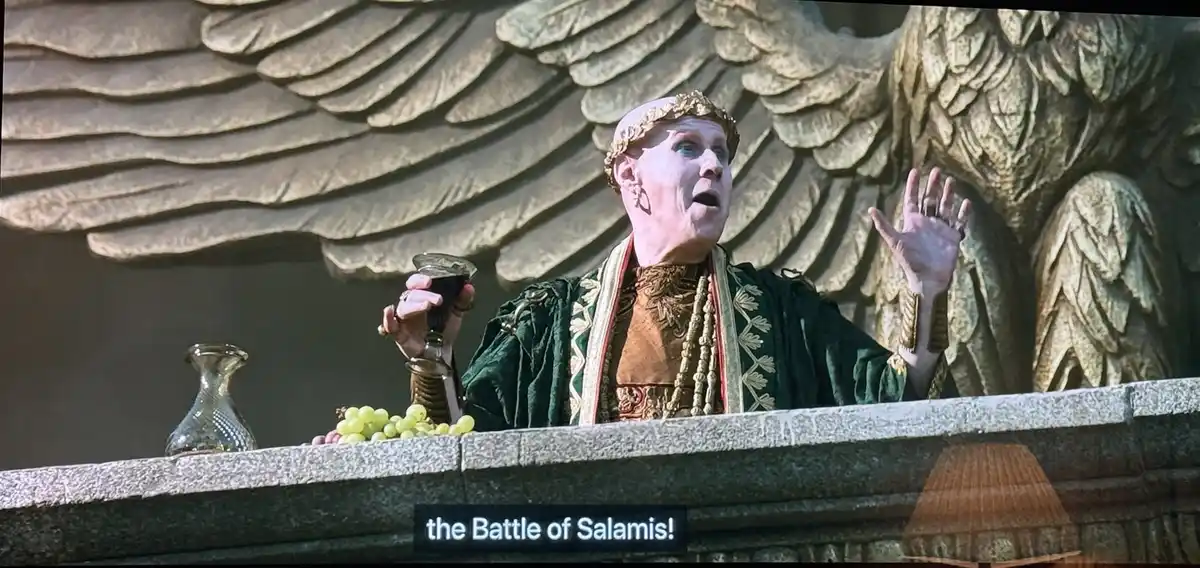 A bald man in ornate ancient Greek-style robes and a golden crown stands on a stone balcony, holding a goblet of dark liquid…