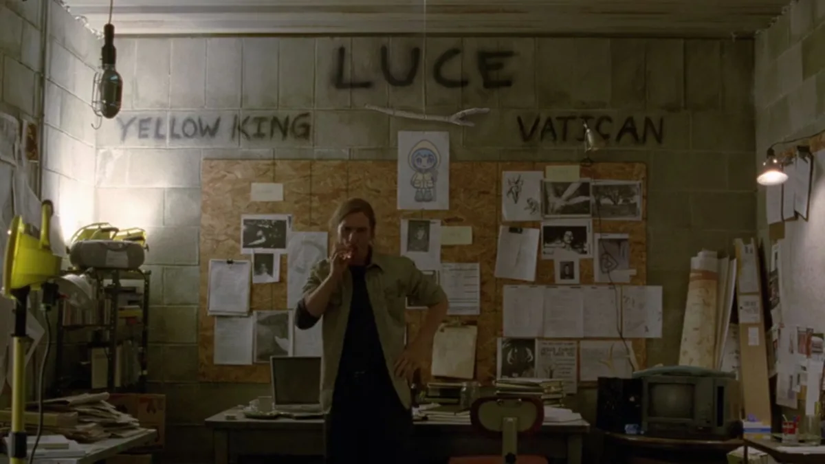A man stands in a cluttered, dimly lit room with cinder block walls, smoking a cigarette. The wall behind him has 'LUCE'…