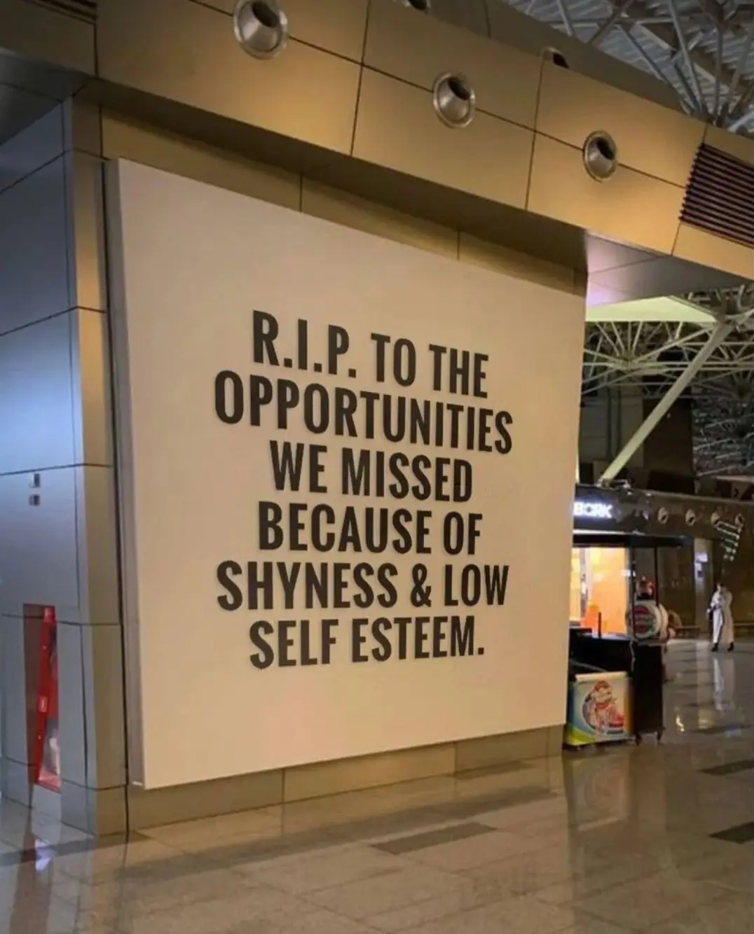A large beige sign in a modern airport terminal reads 'R.I.P. TO THE OPPORTUNITIES WE MISSED BECAUSE OF SHYNESS & LOW SELF…