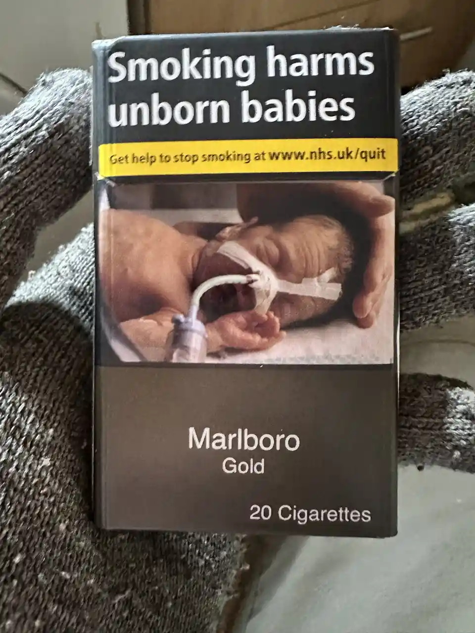 A close-up photo shows a gloved hand holding a standard Marlboro Gold cigarette pack. The pack features the UK government's…