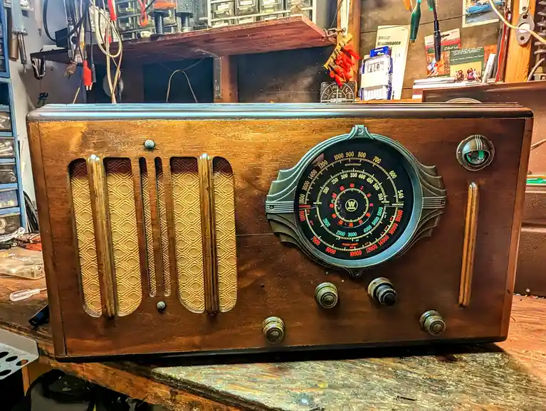 A vintage wooden radio with a large dial and speaker grille sits on a cluttered workbench surrounded by tools and parts. The…