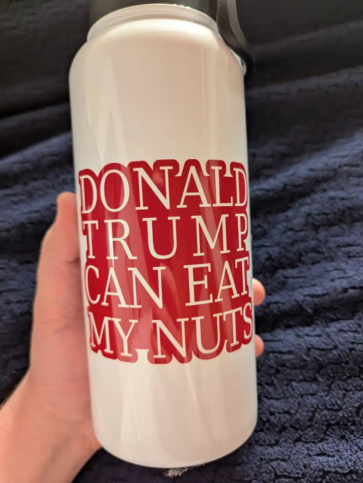 A hand holds a white water bottle with bold red text that reads 'DONALD TRUMP CAN EAT MY NUTS'. The bottle has a black lid…