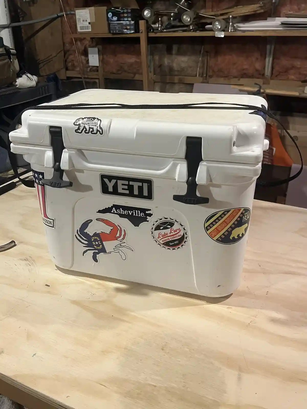 A white Yeti cooler sits on a wooden table in a cluttered garage, covered in various stickers including 'Asheville', 'Lake…