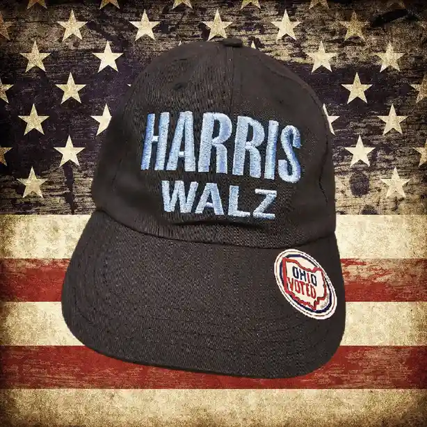 A black baseball cap with 'HARRIS WALZ' embroidered in light blue is centered against a distressed American flag background…