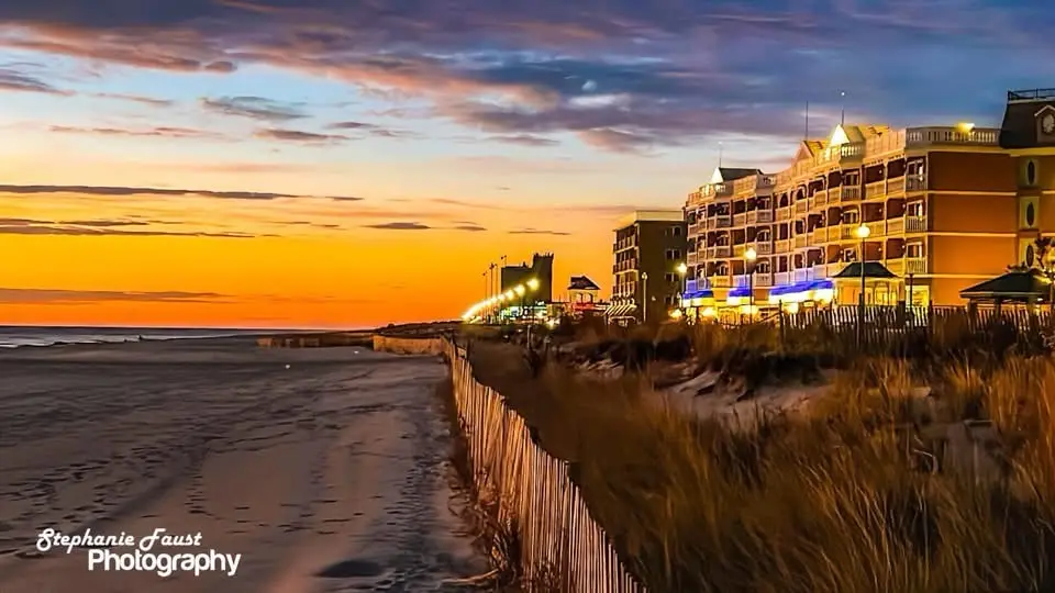 A scenic beach sunset photo with a multi-story hotel lit up on the right, a wooden fence and dunes in the foreground, and a…