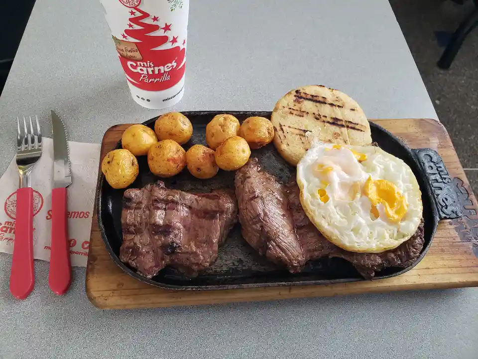 A sizzling cast-iron plate from 'mis Carnes Parrilla' features two grilled steaks, a fried egg, roasted potatoes, and a…