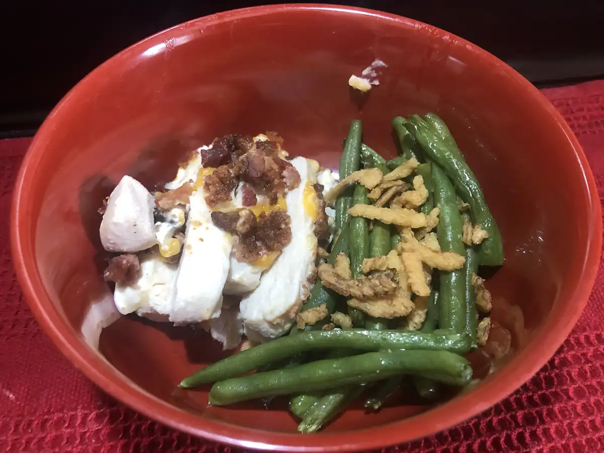 A red bowl contains a serving of sliced chicken topped with bacon and cheese, alongside green beans garnished with fried…