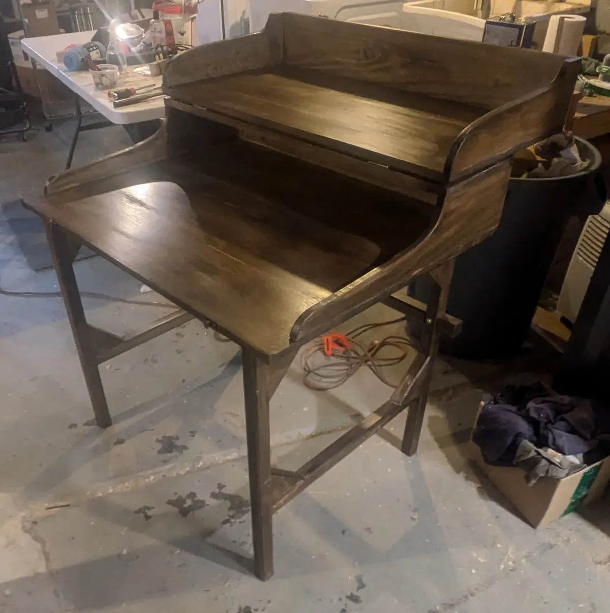 A dark wooden, vintage-style school desk with a raised back and slanted writing surface sits in a cluttered workshop. The…