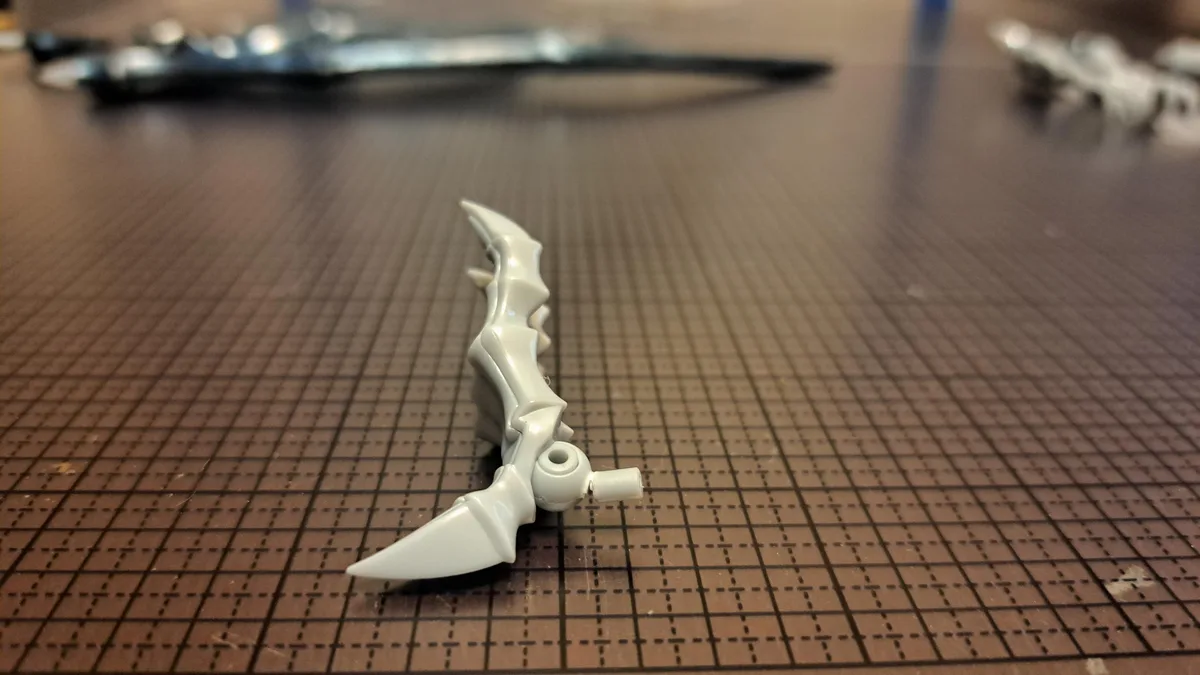 A close-up photo shows a white, plastic, ornate weapon part, likely from a model kit, resting on a brown grid-patterned work…
