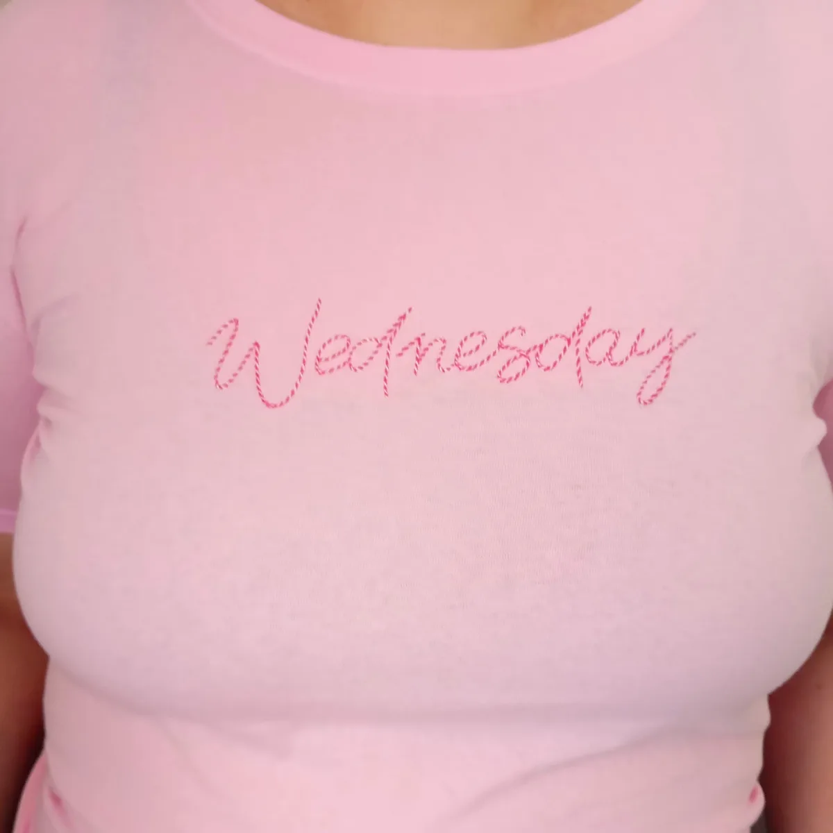 A close-up of a person wearing a light pink t-shirt with the word 'Wednesday' embroidered in red thread across the chest in…