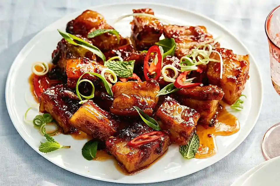 A close-up photo shows a white plate piled high with glistening, braised pork belly pieces coated in a rich, dark sauce. The…
