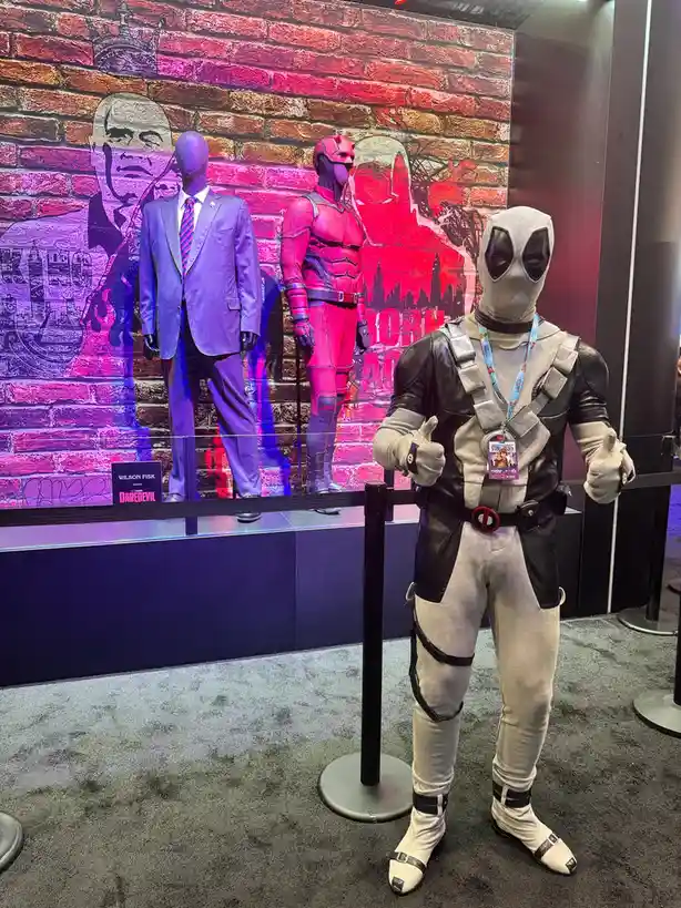 A mannequin dressed as Deadpool stands in front of a display case with two other mannequins, one in a purple suit labeled…